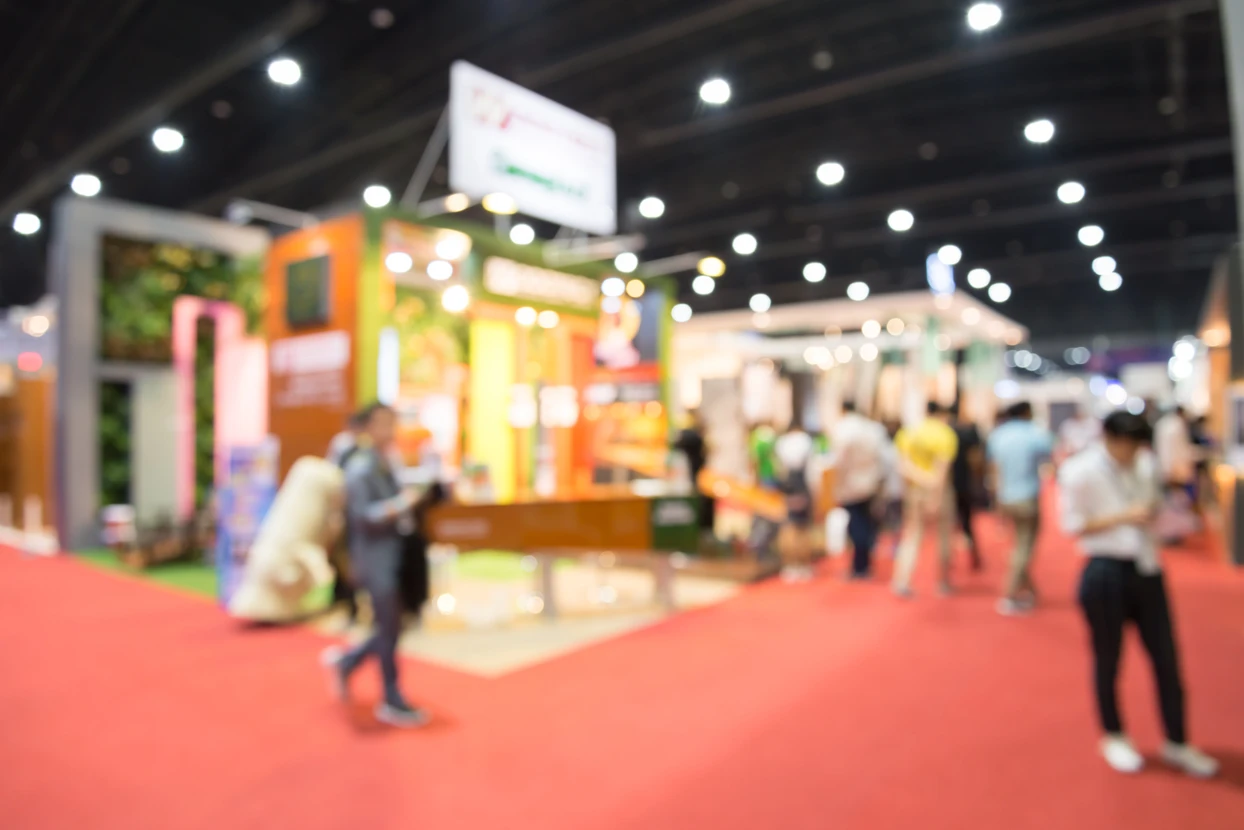 Salon professionnel avec stands d'exposition colorés, visiteurs circulant sur moquette rouge, éclairage LED plafonnier dans hall d'exposition